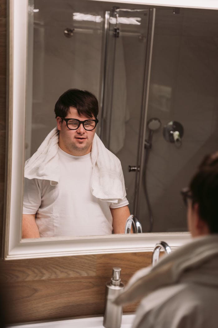 Man with Down Syndrome wearing glasses and towel in bathroom mirror reflection.